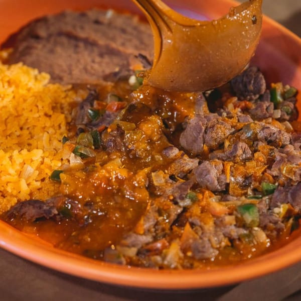 A spoon pouring red sauce over a Mexican dish with meat, rice, and refried beans.