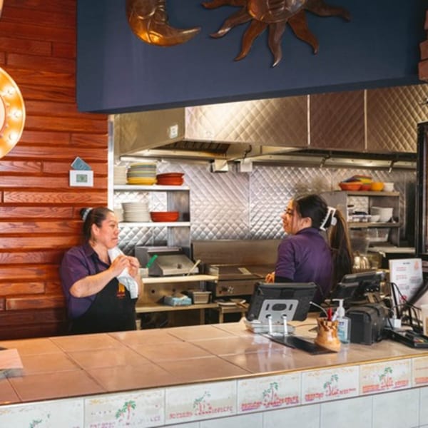 Two women working behind the counter in a Mexican restaurant kitchen with decorative sun and moon.