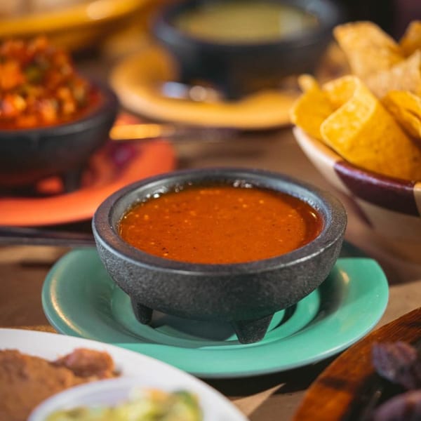 Close-up of a molcajete bowl filled with red salsa, with chips and other salsas blurred in the background.
