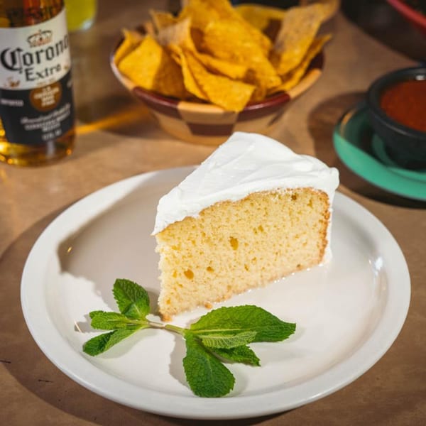 Slice of Tres Leches cake with mint on a white plate, with chips, salsa, and Corona in the background.