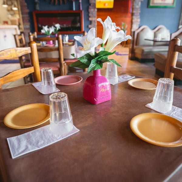 A dining table set with plates, glasses, and a pink Patron bottle vase with white lilies at La Playita.